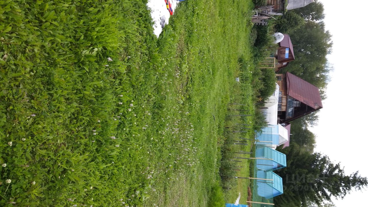 На фотографии представлено просторное зеленое пространство с ухоженной травой и небольшими цветущими растениями. Видны элементы ландшафтного дизайна, такие как деревянные конструкции и заборы. Вдали расположены дома с крышей красного цвета и голубыми элементами, что создает гармоничное сочетание с природой. Участок выглядит идеально подходящим для отдыха на свежем воздухе. Структурные особенности включают в себя разнообразные постройки, которые добавляют уют и индивидуальность этому месту. Общая атмосфера спокойствия и уединения подчеркивает природное окружение.