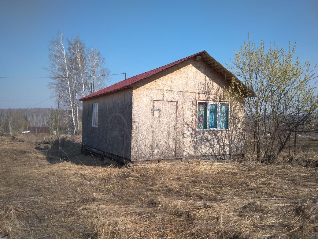 Здание выполнено в простом архитектурном стиле с использованием древесно-стружечных плит. Структурные особенности включают двускатную крышу с красной металлочерепицей и окна с простыми деревянными рамами. Видна дверь, расположенная на передней стороне, и несколько окон, обеспечивающих естественное освещение. Цвета фасада нейтральные, с преобладанием светлых и коричневых оттенков, что создает гармоничный вид. Отсутствие сложных отделочных материалов придает дому скромный и практичный облик, подходящий для загородного проживания или дачи.