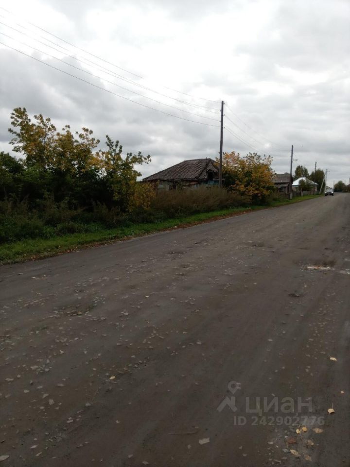 На фотографии видна дорога с асфальтовым покрытием, окруженная зелеными насаждениями. Слева расположены несколько деревьев с желтыми и зелеными листьями, создающими осеннюю атмосферу. На заднем плане виден дом с крышей из черепицы, который имеет простую архитектурную форму и требует ремонта. Стены дома окрашены в светлый цвет, а окна обрамлены деревянными рамами. Вдоль дороги стоят электрические столбы, что указывает на наличие коммуникаций. Общая атмосфера места спокойная и уединенная, с характером загородной жизни.
