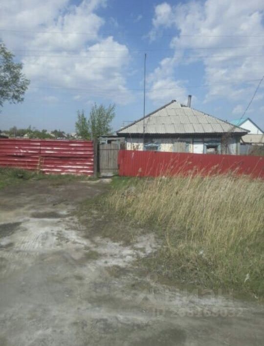 На фотографии представлено здание с простой архитектурой и крышей с наклоном. Стены дома покрыты светлой краской, а окна имеют деревянные рамы. Входная дверь находится сбоку, рядом с ней расположен забор из металлических листов красного цвета. Перед домом видна неухоженная территория с травой и грязным грунтом, что создает ощущение запущенности. Общее оформление и отделка здания простые, без излишеств, что подчеркивает его функциональность. Вокруг дома растут кустарники, добавляющие немного зелени к общему виду.