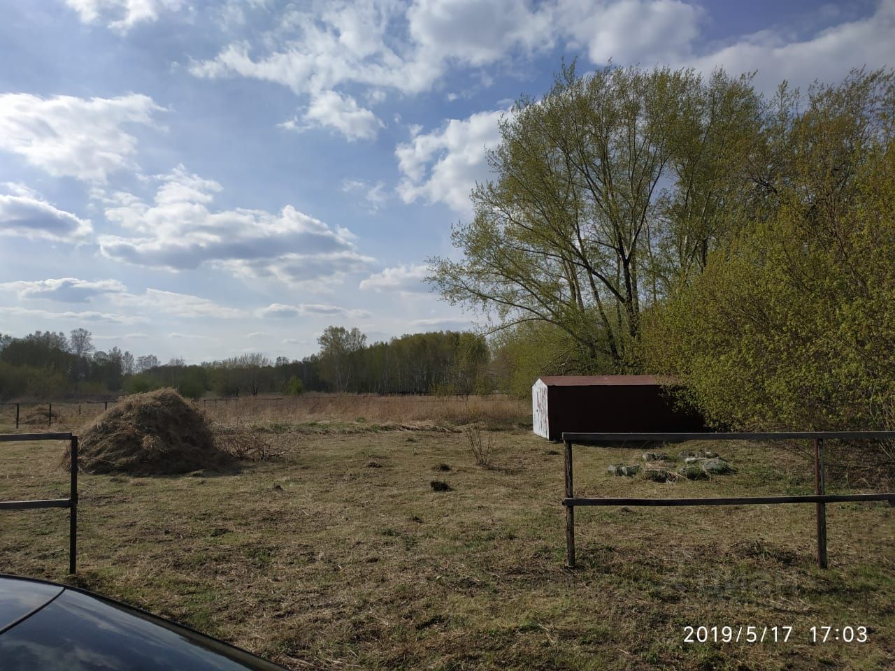 На фотографии видно открытое пространство с ухоженной травянистой местностью. В центре кадра находится куча сена, а на заднем плане виден небольшой сарай красного цвета с белыми элементами. Около сарая расположены камни, а по краям видны металлические ограждения. Слева от изображения можно заметить часть автомобиля. Вдали виднеются деревья, создающие ощущение уединения и природной гармонии. Цвета в основном теплые, с преобладанием зелёного и коричневого, что подчеркивает сельский характер данного участка.