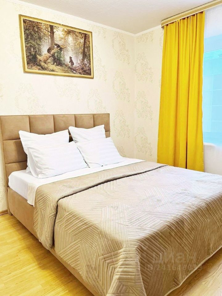 Уютная спальня с двуспальной кроватью, светлыми обоями и желтыми шторами. Картина на стене добавляет атмосферу.