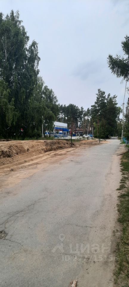 На изображении видна улица с асфальтированным покрытием, окруженная зелеными деревьями и кустарниками. Дорожное полотно имеет небольшие повреждения и трещины, что указывает на необходимость ремонта. С одной стороны улицы расположены столбы освещения, а также видна часть здания с яркой окраской, вероятно, это коммерческое или административное здание. Вдоль дороги заметны участки с выемками грунта, что может свидетельствовать о проведении строительных или ремонтных работ. Общая атмосфера пространства спокойная и уединенная, создавая ощущение пригородной местности.