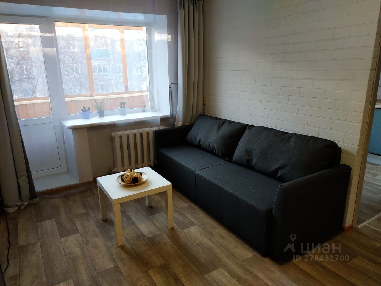 Интерьер комнаты выполнен в современном стиле с акцентом на минимализм. На переднем плане расположен угловой диван темного цвета, который гармонирует с деревянным полом. Белый столик с декоративными элементами, такими как корзины, добавляет уюта. Окно с белыми рамами и прозрачными шторами обеспечивает естественное освещение и открывает вид на балкон, где находятся растения. Стены окрашены в светлый цвет, создавая ощущение простора и светлоты. Радиатор отопления под окном дополняет функциональность пространства, а общая цветовая палитра создает спокойную атмосферу.