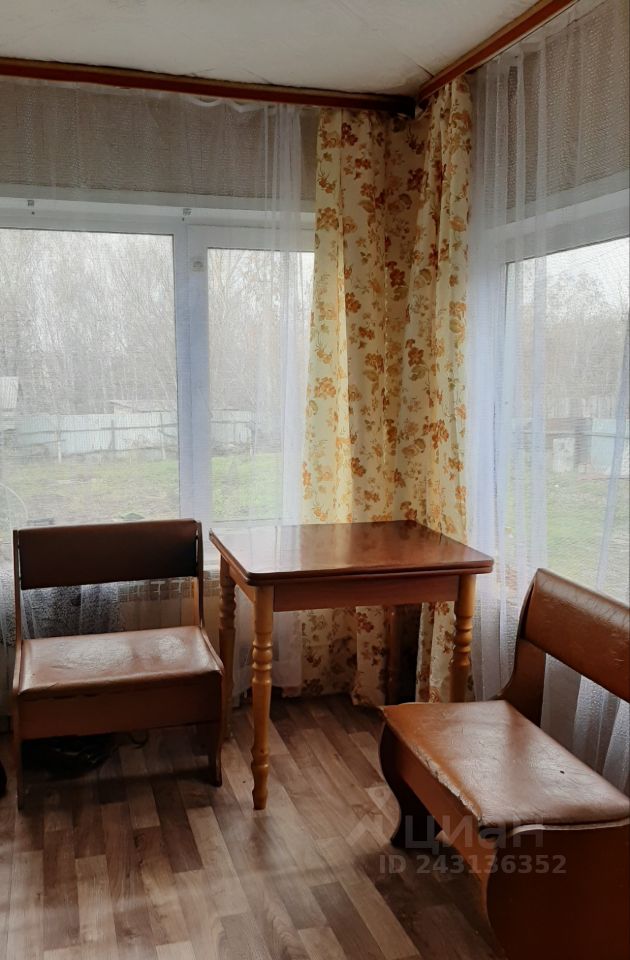Уютное помещение с большими окнами, обрамленными легкими занавесками. Стены оформлены в светлых тонах, а в углу видна яркая цветочная штора. На полу уложен ламинат, создающий теплую атмосферу. В центре стоит деревянный стол с резными ножками, окруженный двумя стульями с кожаной обивкой. Мебель имеет простые, но функциональные формы, что подчеркивает практичность пространства. Комната выглядит светлой и приветливой, идеально подходящей для отдыха или общения.