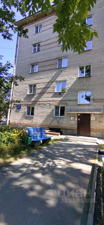Фасад здания выполнен в простом, функциональном стиле с использованием светлого кирпича и горизонтальных полос. На первом этаже расположены входные двери и окна, оформленные в стандартном формате. Входная группа имеет небольшую лестницу и площадку, где установлены деревянные скамейки. Цветовая палитра включает светлые и нейтральные оттенки, что создает уютное впечатление. Вокруг видны зеленые насаждения и цветы, что добавляет живописности. Основное внимание уделено практичности и доступности, что характерно для жилых зданий данного типа.
