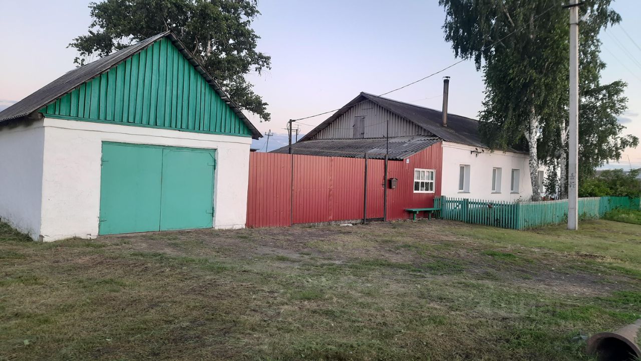 Здание выполнено в простом, функциональном архитектурном стиле с элементами традиционного дизайна. Видны два строения: одно с зеленой крышей и белыми стенами, другое с серой крышей и красными панелями. Структура включает гараж с большими зелеными воротами и жилое помещение с окнами, оформленными в современном стиле. Основные цвета — белый, зеленый и красный, что создает контраст. Отделка стен простая, без лишних украшений, подчеркивающая практичность. Пространство вокруг аккуратно оформлено, с зеленым забором и небольшой скамейкой.