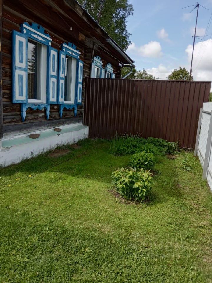На фотографии представлено здание в традиционном русском стиле с характерными деревянными элементами. Окна оформлены яркими синими наличниками, что придаёт фасаду выразительность. Стена здания выполнена из тёмного дерева, что создаёт контраст с белыми рамами окон. Внизу под окнами видны каменные подоконники. Перед зданием расположен ухоженный участок с зелёной травой и небольшими кустами, что добавляет уюта. Забор из коричневого металла ограничивает пространство, создавая приватность. Общая атмосфера вызывает ощущение традиционного русского быта.