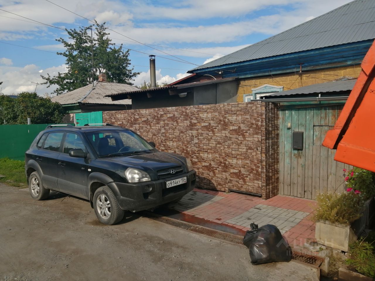 На фотографии виден участок с асфальтированной парковкой для автомобиля и ограждением из искусственного камня. Стена выполнена в коричневых тонах, создавая эффект натурального камня. Дверь гаража из деревянных панелей с зелеными ставнями придает пространству уютный вид. Пол выложен плиткой, сочетание серых и красных оттенков. На переднем плане находится черный внедорожник, который занимает часть парковки. Рядом с ним видна мусорная袋 и клумба с цветами, добавляющая яркие акценты в общую композицию.