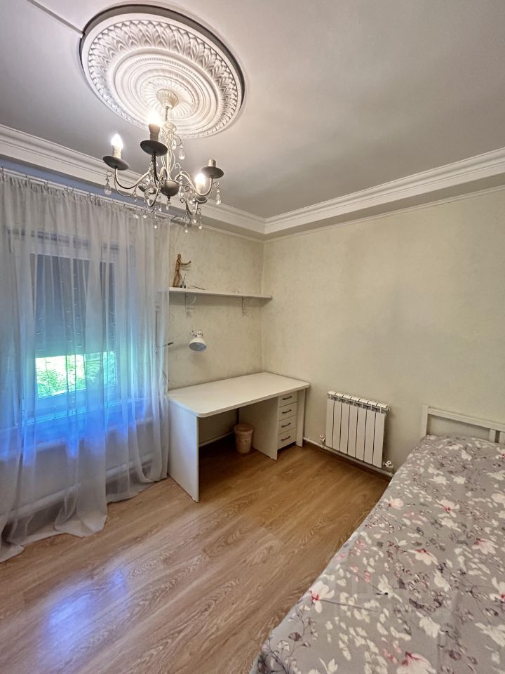Комната оформлена в современном стиле с элементами классического дизайна. На потолке расположена декоративная лепнина и стильная люстра. Стены окрашены в светлый цвет, создавая ощущение простора. У окна висят легкие прозрачные шторы, пропускающие естественный свет. В углу находится письменный стол с настольной лампой, рядом с ним расположен обогреватель. Пол выполнен из светлого laminata, что придает комнате уют. Кровать с цветочным покрывалом добавляет теплоты и уюта, а минималистичная мебель подчеркивает функциональность пространства.