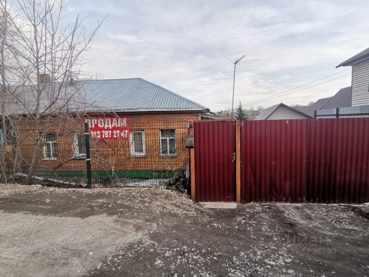 На фотографии представлено здание с кирпичной фасадом в традиционном архитектурном стиле. Видны окна с простыми рамами и металлическая ограда, разделяющая участок. Крыша имеет скатную форму с металлическим покрытием. Входные ворота выполнены из металлического профиля, окрашенного в бордовый цвет, что создает контраст с кирпичной стеной. На стене здания размещена вывеска с надписью 'Продаем', что указывает на его коммерческое назначение. В целом, пространство выглядит ухоженным, но требует некоторого внимания к деталям.