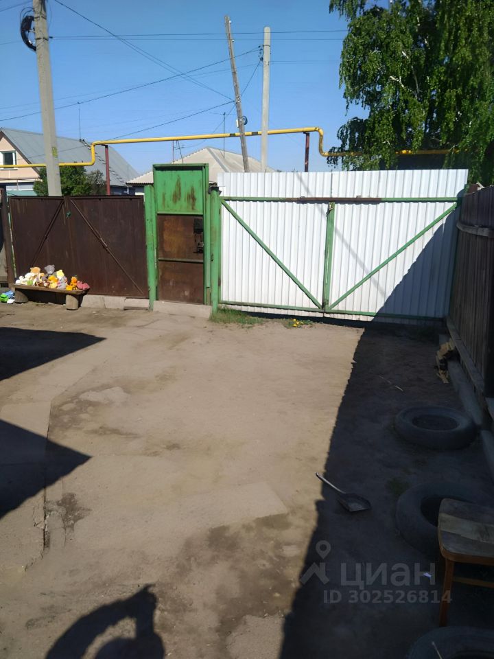 На фотографии виден участок с простым асфальтовым покрытием и песчаной поверхностью. Справа расположена деревянная скамейка, а рядом лежат покрышки. Основной элемент — это ворота, выполненные из металлических и деревянных конструкций, окрашенные в зеленый цвет. Ворота открываются на улицу, создавая доступ к участку. Вдали можно увидеть другие строения, что указывает на жилую застройку. Цвета в основном нейтральные, с акцентами зеленого и коричневого. Пространство выглядит незагроможденным и функциональным.