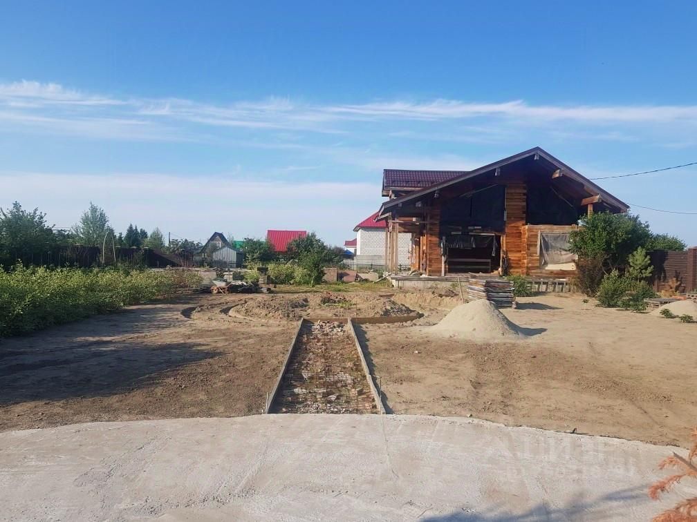 На фотографии представлено деревянное строение в загородном стиле с крышей из металлочерепицы. Архитектурные элементы включают открытые террасы и большие окна, обеспечивающие естественное освещение. Внешняя отделка выполнена из бруса, что придает дому уютный вид. Перед зданием видна территория с подготовленной площадкой, на которой находятся строительные материалы и песок. Ландшафт вокруг дома включает зелёные насаждения и участки с подготовленной почвой, что создаёт возможность для дальнейшего озеленения и благоустройства. Цветовая палитра включает натуральные древесные оттенки и нейтральные тона.