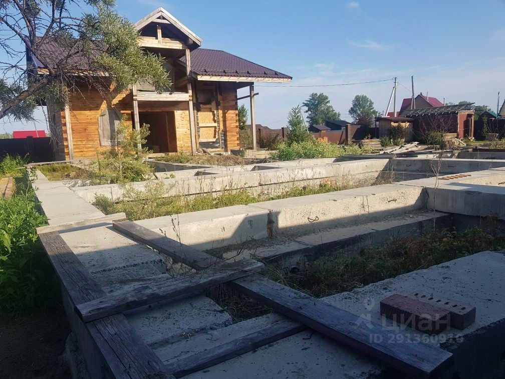 Архитектурный стиль здания выполнен в загородном деревянном стиле с элементами традиционного дизайна. Структурные особенности включают в себя деревянные балки и крыши с наклоном, покрытые черепицей. Видна площадь, на которой расположены фундаменты, окруженные дикими растениями. Основные характеристики пространства – это открытая планировка, предполагающая возможность создания различных комнат. Материалы включают дерево для стен и бетон для фундамента, а цвета варьируются от светлого дерева до серых бетонных конструкций, что создает контраст в отделке.