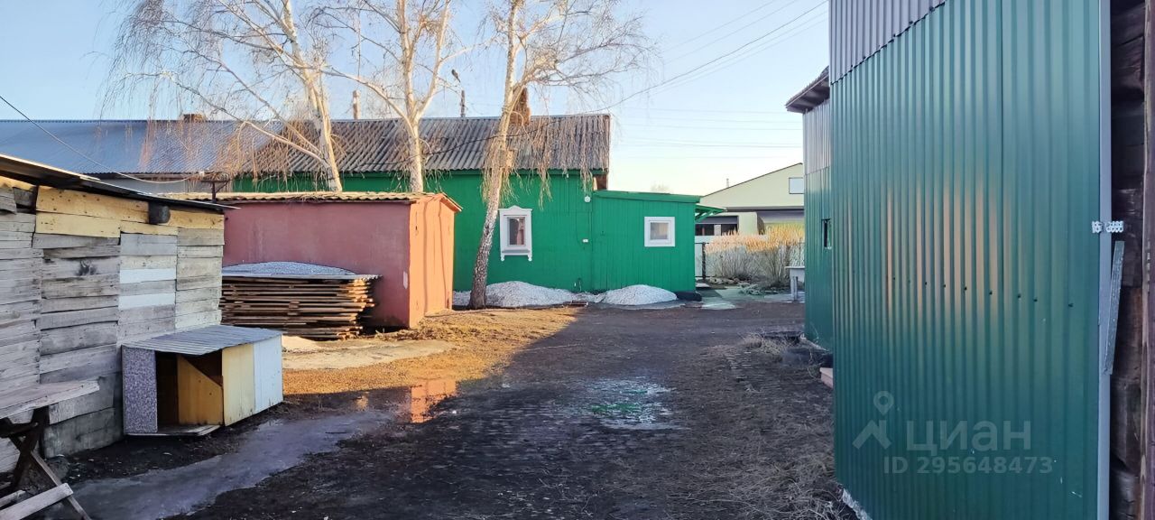 На фотографии представлена территория с несколькими постройками, выполненными в простом функциональном стиле. Основные элементы конструкции включают деревянные и металлические стены, а также крыши с наклоном. Зелёный и красный цвета зданий создают контрастный вид. В центре видно место для хранения материалов, таких как деревянные доски. Пол вымощен гравием, а участок вокруг имеет ухоженный вид. Пространство выглядит организованным, с чётким разделением на зоны для различных нужд. Основные отделочные материалы включают дерево и металл, что придаёт постройкам практичность и долговечность.