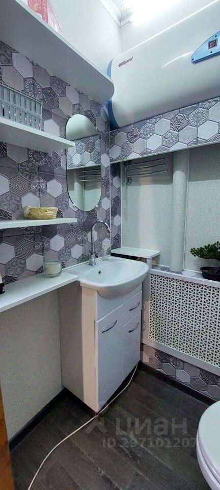 В ванной комнате представлен современный дизайн с геометрической плиткой в виде шестиугольников, что придаёт пространству стильный вид. Основные элементы включают компактную раковину с тумбой, выполненной в белом цвете, и зеркало, расположенное над раковиной. Стены отделаны плиткой с узором, а пол покрыт тёмным ламинатом, создающим контраст. На открытых полках размещены аксессуары, добавляющие функциональность. В комнате также виден водонагреватель, установленный на стене, что свидетельствует о современных удобствах. Общая цветовая палитра включает белые и серые тона, создавая ощущение чистоты и свежести.