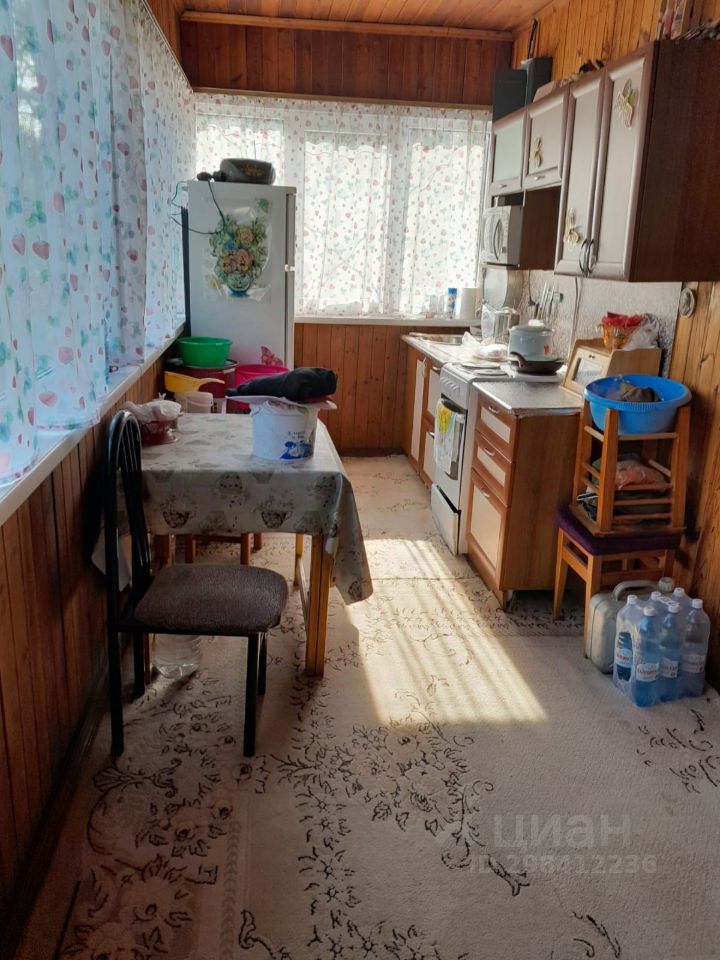 Кухня с деревянными стенами и потолком, оформленная в традиционном стиле. На стенах расположены шкафы с деревянными фасадами, а также бытовая техника, включая плиту и холодильник. В центре комнаты стоит стол с скатертью, окруженный стульями. Пол устлан ковром с узором, а на столе находятся предметы кухонной утвари. Окна обрамлены легкими занавесками с цветочным рисунком, что придаёт помещению уют. В углу видны пластиковые контейнеры и бутылки с водой, что указывает на активное использование пространства.