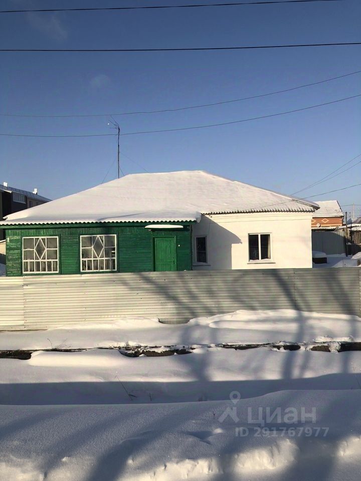 На фотографии представлено одноэтажное здание с характерным архитектурным стилем. Фасад выполнен в двух цветах: зеленом и белом, что создает контраст. Окна имеют традиционные рамы с несколькими секциями, а входная дверь выделяется зеленым цветом. Крыша имеет простой, но функциональный дизайн, покрыта снегом. Стены выглядят прочными, а забор из металлических панелей добавляет уединения. Внутреннее пространство, вероятно, светлое, благодаря большим окнам, которые обеспечивают естественное освещение. Общая атмосфера здания вызывает ощущение уюта и простоты.