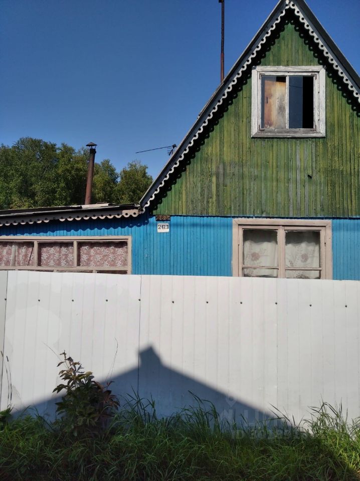Дом в народном архитектурном стиле с характерными элементами, такими как треугольная крыша и деревянная отделка. Стены окрашены в яркие цвета: зелёный и синий, что придаёт зданию уникальный вид. Окна с деревянными рамами и занавесками, частично закрытыми. Видны элементы традиционного дизайна, такие как карнизы и резные детали. Структура дома демонстрирует простоту и функциональность, с акцентом на природные материалы. Фасад окружён белым забором, который контрастирует с яркими цветами стен, создавая уютную атмосферу.