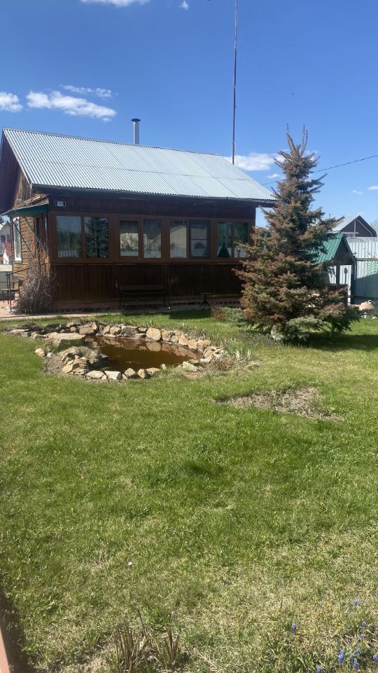Деревянный дом в загородном стиле с металлической крышей и большими окнами, обеспечивающими хорошее освещение. Стены отделаны натуральным деревом, что придает уютный вид. Перед домом расположен небольшой пруд, окруженный каменной окантовкой. На переднем плане ухоженный зеленый газон, который создает приятную атмосферу. Вокруг растут хвойные деревья, добавляющие природный элемент. Общая цветовая гамма включает теплые коричневые и зеленые тона, что подчеркивает гармонию с природой.