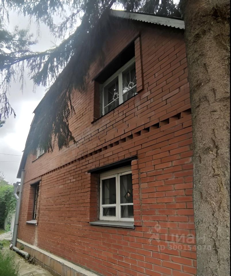 Стена дома выполнена из красного кирпича с характерной текстурой и горизонтальными линиями. Окна имеют белые рамы и решетки, что придает зданию классический вид. Крыша с небольшим свесом и металлическим покрытием завершает архитектурный стиль. Структурные элементы включают вертикальные и горизонтальные линии, создающие визуальный ритм. На фасаде видны элементы отделки, такие как простые карнизы и наличники вокруг окон. Общая цветовая гамма теплых тонов кирпича и светлых рам окон гармонирует с дизайном, создавая уютное впечатление.