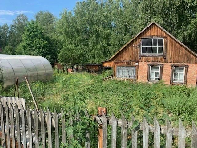 Деревянный дом с кирпичными элементами, выполненный в традиционном русском стиле, имеет двускатную крышу и большие окна. Стены частично облицованы кирпичом, что придаёт зданию устойчивый и теплый вид. На переднем плане виден участок с высокой травой и забором из деревянных досок. Слева расположена теплица с прозрачным покрытием, что указывает на возможность садоводства. Основные цвета — теплые оттенки дерева и кирпича, создающие уютную атмосферу. Архитектурные детали, такие как резные наличники на окнах, подчеркивают народный стиль.