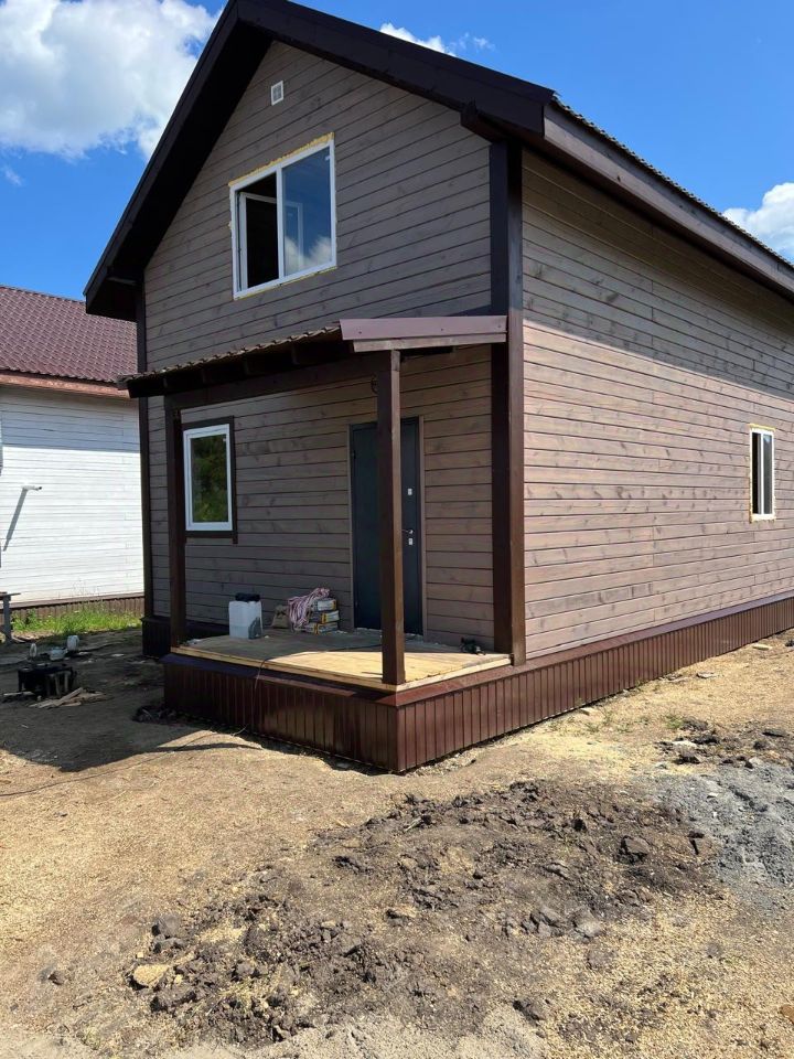 Архитектурный стиль здания представлен в виде современного дома с простыми линиями и деревянной отделкой. Структурные особенности включают двускатную крышу и просторную террасу с перилами. На переднем плане виден вход с дверью и окнами, которые обеспечивают естественное освещение. Основные характеристики включают использование натурального дерева в отделке, что придаёт дому уютный вид. Цветовая гамма состоит из теплых коричневых оттенков, что создает гармоничную атмосферу. Завершенные отделочные работы придают зданию завершенный и аккуратный вид, подчеркивая его современный дизайн.