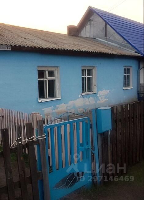 Здание выполнено в простом архитектурном стиле с характерной для загородных домов отделкой. Фасад окрашен в светло-голубой цвет, что придаёт ему свежий вид. Окна с белыми рамами и простыми формами создают симметричный вид. Крыша с наклоном покрыта металлочерепицей синего цвета. Входная зона обрамлена деревянным забором, выполненным из неокрашенных досок, и металлическими воротами, покрашенными в яркий голубой цвет. Общая отделка здания и забор гармонично сочетаются, создавая уютную атмосферу загородного дома.