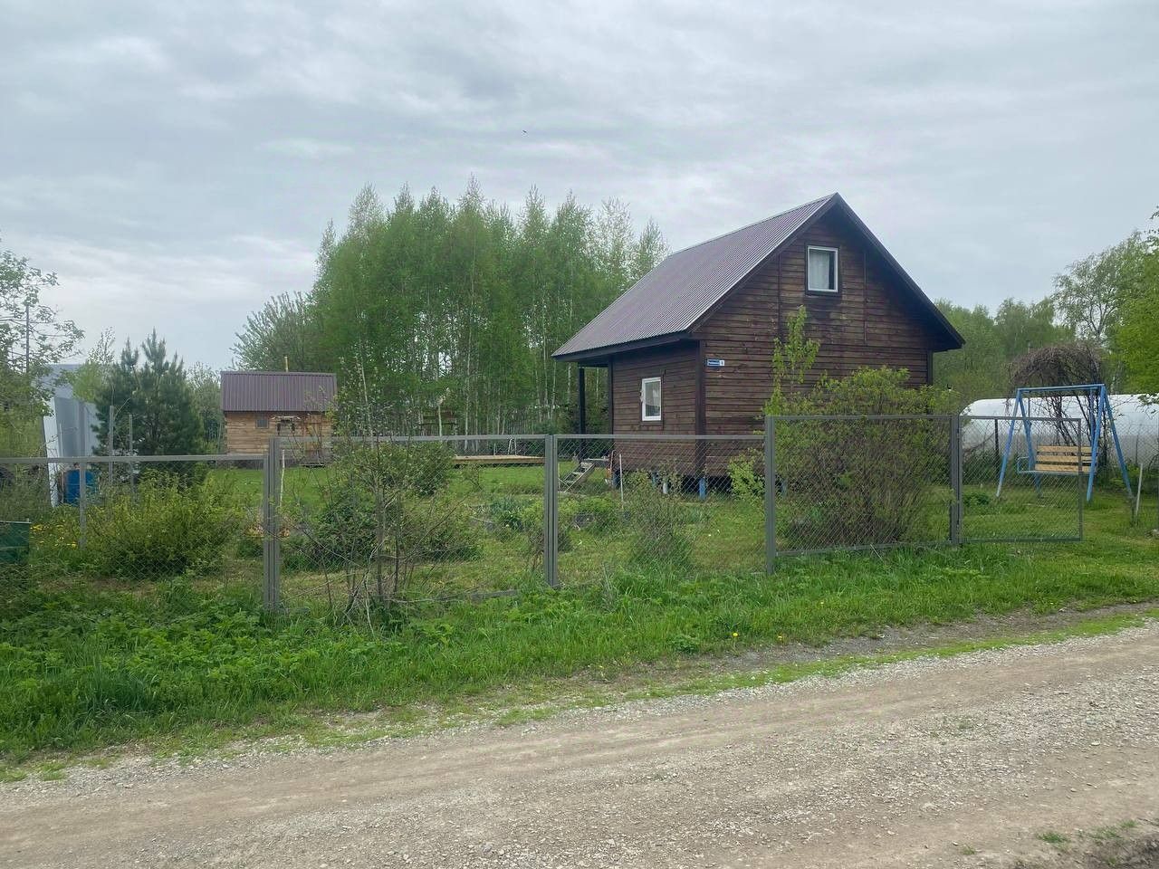 Деревянный дом в загородном стиле с двускатной крышей и небольшими окнами. Стены выполнены из натурального дерева, придающего уютный и теплый вид. Вокруг дома расположен ухоженный участок с зелеными насаждениями и кустарниками. Огороженная территория с металлическим забором создает приватность. На переднем плане виден качельный комплекс, добавляющий элемент отдыха. Цветовая гамма включает естественные древесные оттенки и зеленые тона растительности, создавая гармоничное сочетание с окружающей природой.