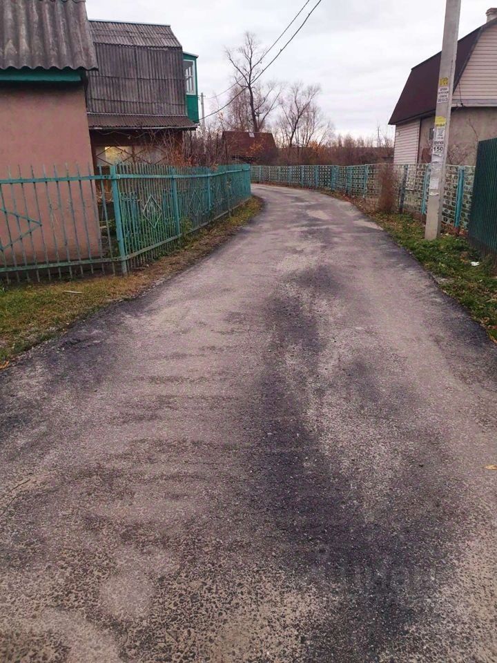 На фотографии видно асфальтированное дорожное покрытие, которое извивается между домами. С обеих сторон дороги расположены металлические заборы зеленого цвета. Один из домов имеет крыши с металлическими покрытиями и балкон, выполненный из стекла и дерева. Дорога окружена участками земли с небольшими кустарниками и травой. Стиль заборов и домов указывает на типичную загородную архитектуру, характерную для дачных участков. Общая атмосфера создает ощущение уединения и спокойствия.