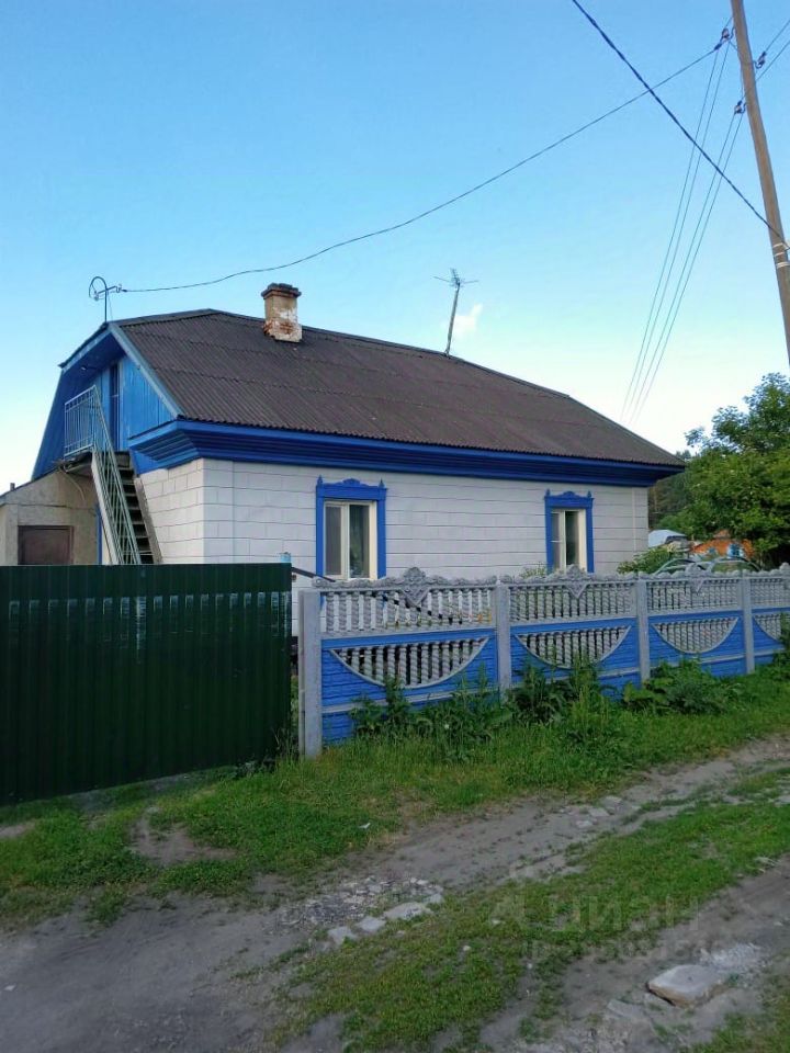 Дом в традиционном русском стиле с белыми стенами и яркими синими декоративными элементами. Крыша с металлической черепицей, что придаёт современный вид. Окна обрамлены синими рамами, что создает контраст с фасадом. Видна лестница, ведущая на второй этаж, что указывает на двухуровневую структуру. Забор из дерева с элементами резьбы добавляет уют и защищенность. Использованы натуральные материалы, создающие гармоничное сочетание с окружающей средой. Цвета фасада теплые и привлекательные, что делает дом заметным и привлекательным.