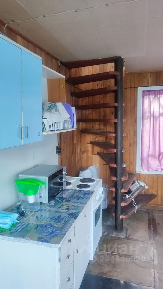 Внутреннее пространство оформлено в простом и функциональном стиле. На стенах видна деревянная отделка, создающая уютную атмосферу. В углу расположена спиральная лестница, ведущая на второй уровень, выполненная из черного металла и дерева. Кухонный уголок представлен белыми шкафчиками и столешницей с ярким рисунком, на которой стоят микроволновая печь и плита. Окно с фиолетовыми занавесками наполняет комнату естественным светом, подчеркивая теплые тона дерева и яркие акценты на кухне.