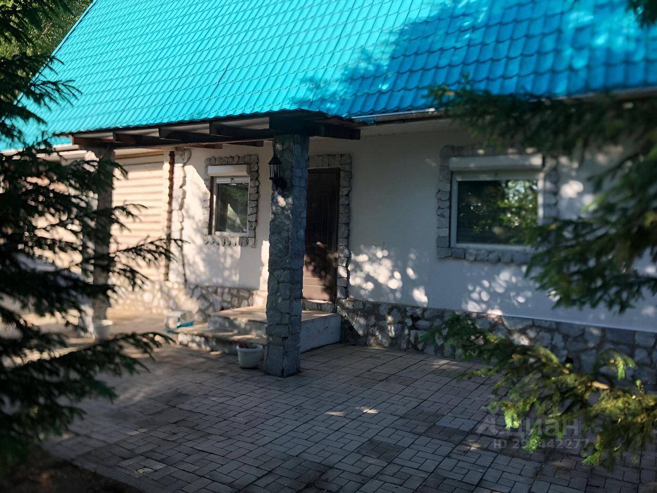 Архитектурный стиль дома сочетает элементы традиционного и современного дизайна. Видны каменные колонны и отделка из природного камня, придающие зданию прочность и уют. Главный вход оформлен деревянной дверью, ведущей на небольшую террасу с плиточным покрытием. Окна имеют простую форму с рамами, выполненными в светлых тонах. Крыша с яркой голубой черепицей привлекает внимание и создает контраст с нейтральными цветами стен. Общая отделка выглядит гармонично и стильно, создавая атмосферу домашнего уюта.