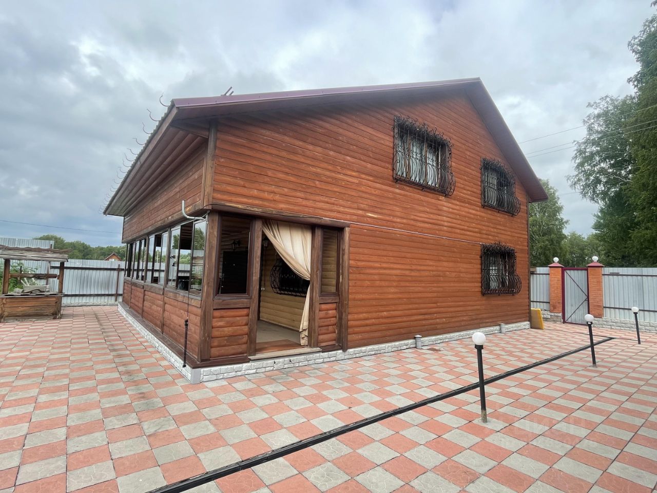Деревянный дом в загородном стиле с отделкой из бруса. Окна украшены металлическими решетками, что придаёт зданию уникальный характер. На фасаде заметны элементы, такие как крыша с небольшим наклоном и терраса с панорамными окнами. Пол вокруг дома выложен плиткой в красно-серых тонах, создавая уютное пространство для отдыха. Стены имеют тёплый коричневый оттенок, а отделка из камня внизу добавляет контраст. Пространство выглядит просторным и хорошо освещённым, с возможностью для создания комфортной зоны для отдыха на свежем воздухе.