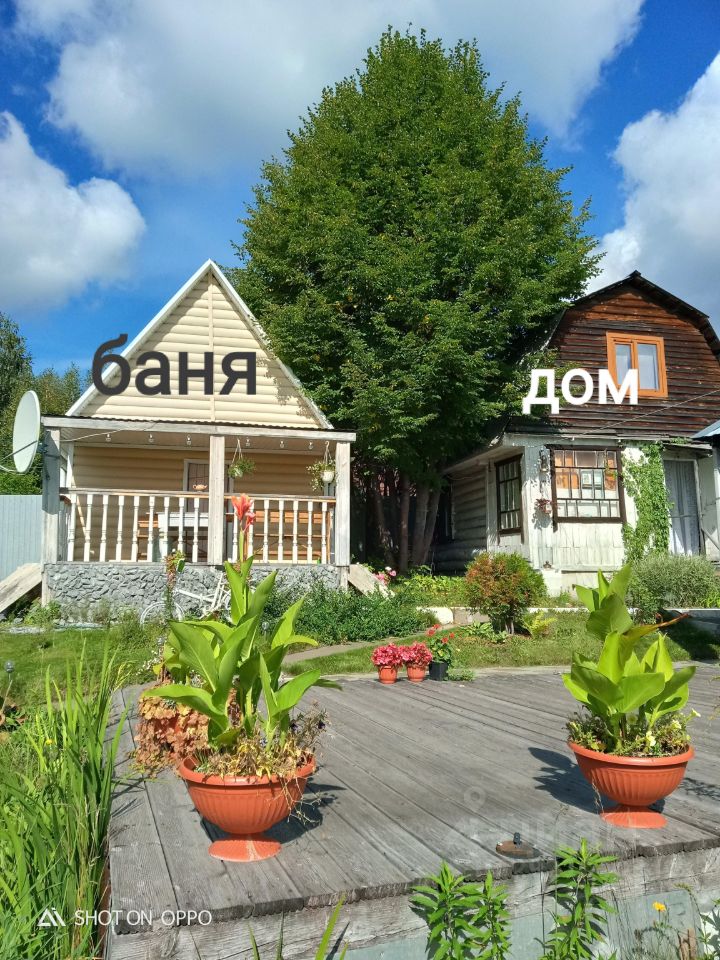 На фотографии изображены два деревянных здания в загородном стиле. Первое здание, баня, имеет светлую обшивку и просторную веранду с колоннами, украшенную цветами. Второе здание, дом, выполнено в более темных тонах с большими окнами, что придает ему уютный вид. Перед зданиями расположена деревянная терраса, на которой стоят горшки с зелеными растениями и цветами, создавая атмосферу уюта и спокойствия. Архитектурные элементы и отделка отражают традиционный загородный стиль, а использование натуральных материалов подчеркивает гармонию с природой.