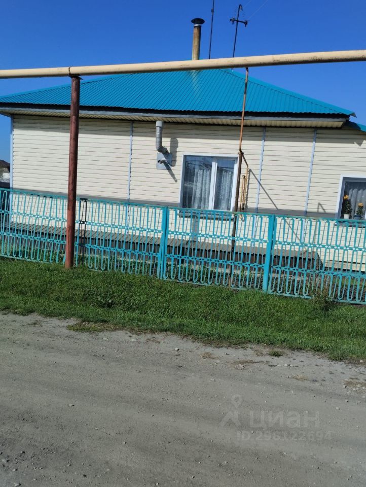Дом в простом архитектурном стиле с двускатной крышей, покрытой голубой металлочерепицей. Фасад выполнен из светлого сайдинга, что придаёт ему современный вид. Окна с белыми рамами и занавесками добавляют уют. Перед домом расположена открытая веранда с перилами, выполненными из металлической сетки. Фасад окружён зелёным забором, который гармонично сочетается с травяным покровом на участке. Структурные элементы, такие как дымоход и водосточные трубы, также заметны, подчеркивая функциональность здания.