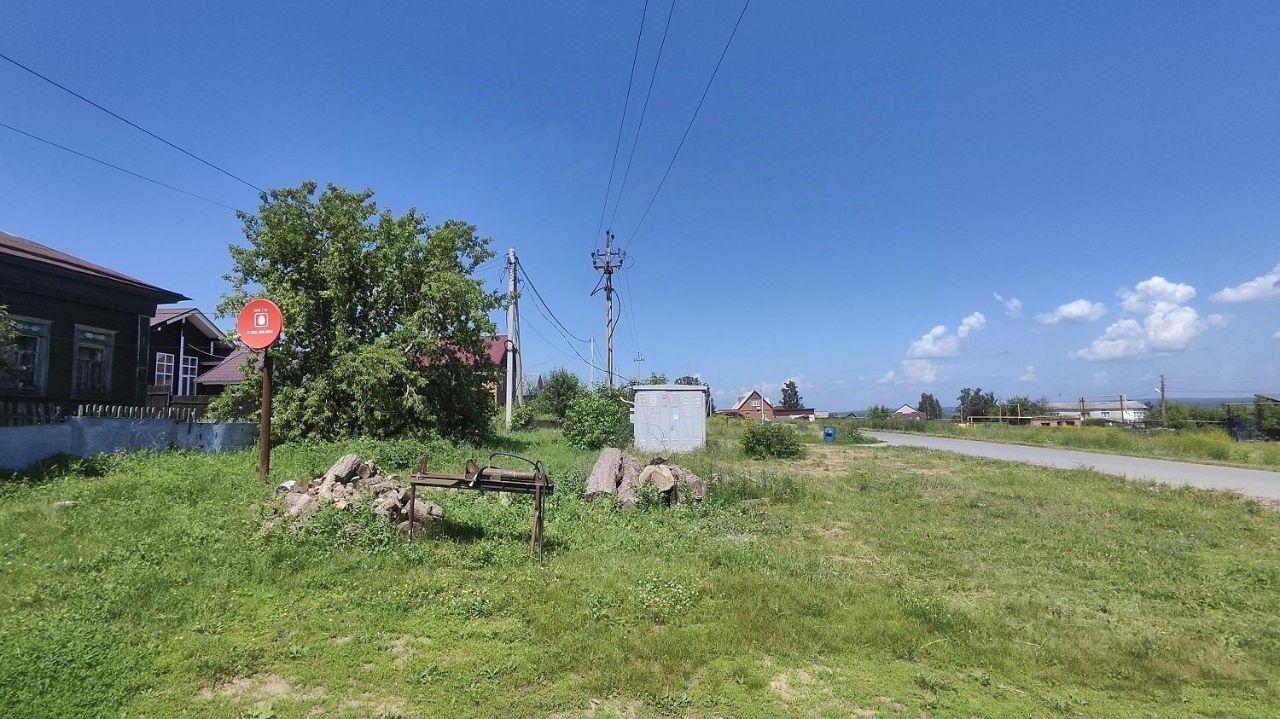 На фото виден участок земли с зелеными травами и кустарниками. Слева расположены деревянные дома с характерными окнами и крышами, покрытыми черепицей. В центре изображения стоит металлическая конструкция, возможно, для хранения дров, рядом с которой видны бревна. На заднем плане можно увидеть электрические столбы и провода, а также небольшой контейнер. Участок выглядит ухоженным, с открытым пространством и аккуратными границами, что создает ощущение спокойствия и уединения.