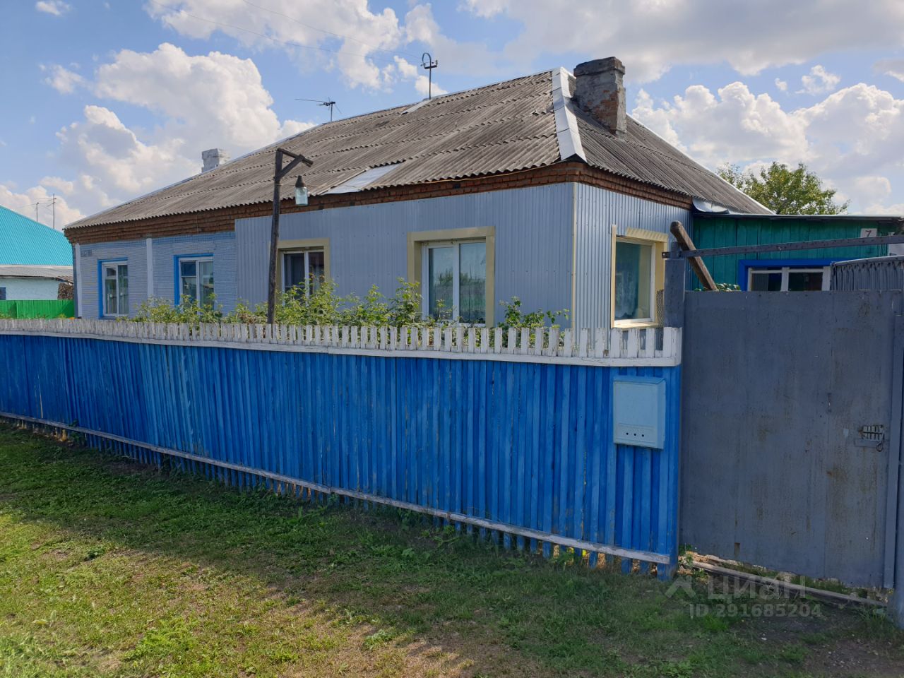 Дом выполнен в простом архитектурном стиле с элементами советского дизайна. Стены окрашены в светло-голубой цвет, а крыша покрыта металлическим листом. Окна имеют деревянные рамы с белыми наличниками, придающими уютный вид. Видно, что дом одноэтажный с небольшим выступом, возможно, террасой. Забор из деревянных досок окрашен в яркий синий цвет, что создает контраст с зеленой растительностью. На переднем плане растут кусты, добавляющие естественности и уюта. Общая атмосфера пространства спокойная и домашняя.