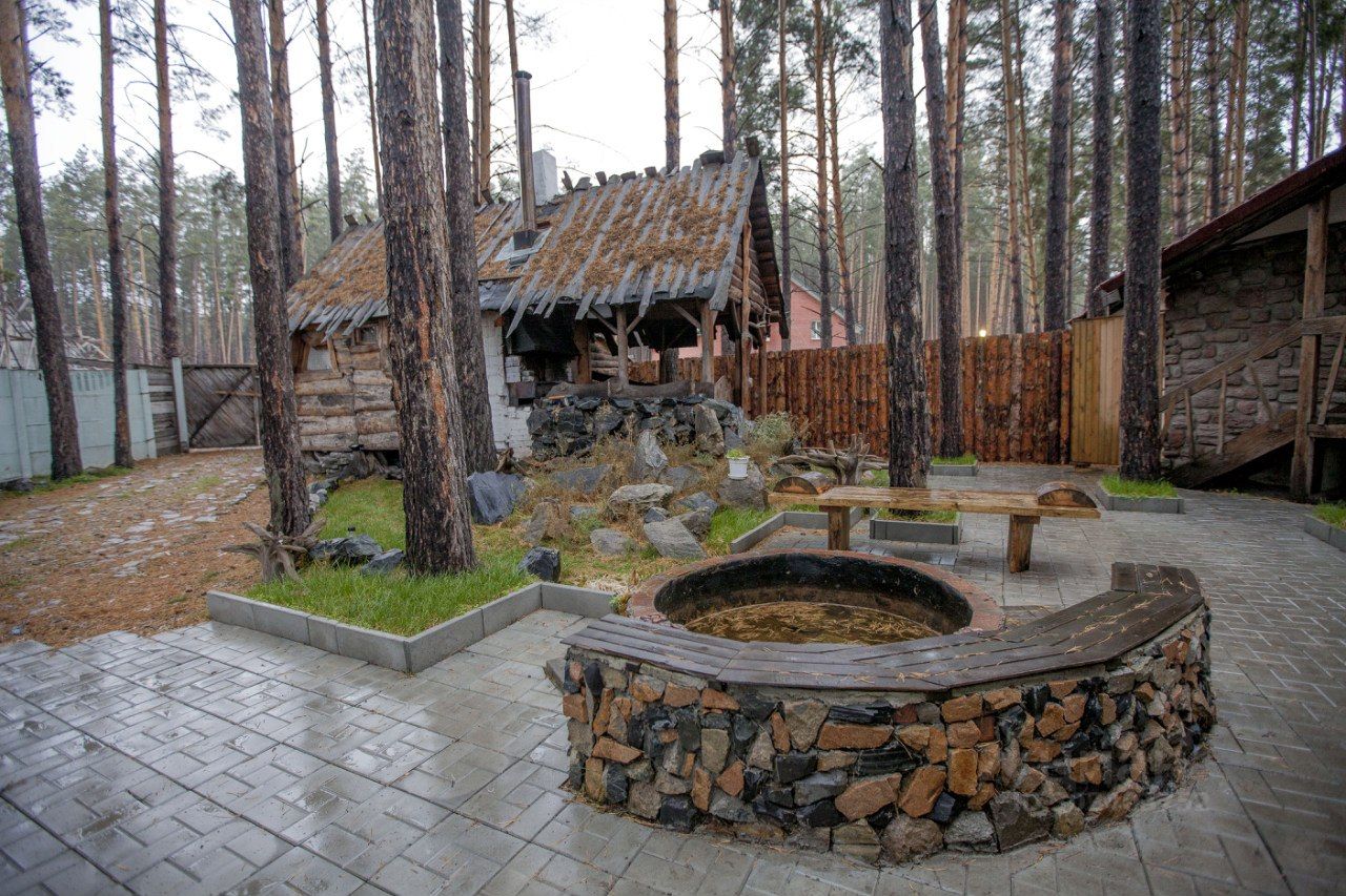 На фото представлен участок с деревянным домом в деревенском стиле. Стены дома выполнены из бревен, крыша покрыта соломой и имеет неформальную форму. В центре участка находится круглая купель, окруженная каменной отделкой и деревянными скамьями. Пол выложен плиткой, а вокруг расположены зеленые зоны с травой и небольшими камнями. Вдалеке видны высокие сосны, создающие уединенную атмосферу. Забор из деревянных досок обрамляет пространство, добавляя уюта и защищенности.