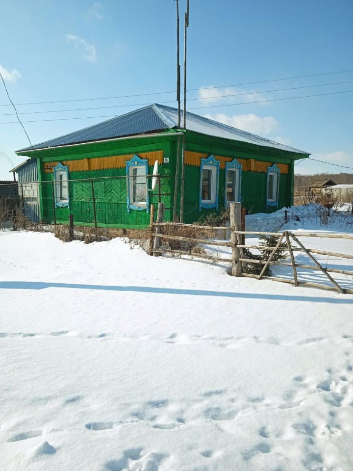 Деревянный дом в традиционном русском стиле, окрашенный в яркий зеленый цвет с желтыми и голубыми элементами. Стены отделаны деревянными панелями с резьбой, придающими зданию уникальный характер. Окна имеют декоративные рамки, выделяющиеся на фоне основного цвета. Крыша двускатная, покрытая металлочерепицей. Перед домом находится деревянный забор, добавляющий уюта. Видно, что вокруг много снега, что подчеркивает зимнюю атмосферу. Архитектурные детали и яркие цвета делают этот дом привлекательным и характерным для русской деревенской архитектуры.