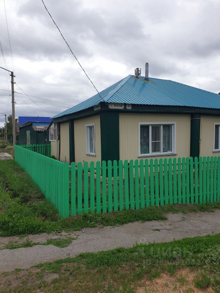 Дом в стиле дачи с округлыми формами и яркой голубой крышей. Стены выполнены из светлого сайдинга, дополненные зелеными вертикальными панелями. Окна с белыми рамами обеспечивают естественное освещение. Вокруг дома установлен зеленый деревянный забор, который создает уютную атмосферу. Дорога перед домом вымощена, а участок окружен зелеными насаждениями. Структурные элементы, такие как дымоход и вентиляция, также видны на крыше. Общая композиция создает гармоничное сочетание цветов и форм, подчеркивающее дачный стиль.