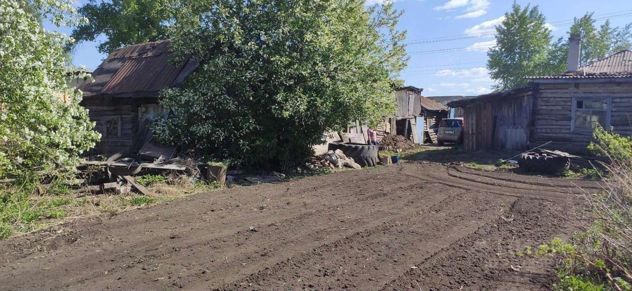 На фотографии виден участок с несколькими деревянными постройками в традиционном русском стиле. Стены зданий выполнены из бревен, некоторые из них имеют ржавые металлические крыши. Участок окружен зелеными деревьями и кустарниками, создающими уютную атмосферу. На земле видно свежепаханную почву, что указывает на возможное использование участка для сельского хозяйства. Вдалеке можно заметить старый автомобиль и несколько хозяйственных предметов, таких как покрышки и деревянные конструкции, что подчеркивает функциональность этого пространства.