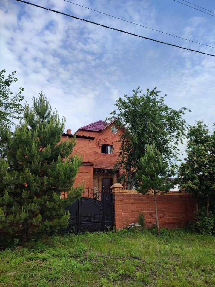 Здание выполнено в кирпичном стиле с элементами классической архитектуры. Фасад украшен симметричными окнами и декоративными деталями. Структурные особенности включают двухэтажную компоновку и мансардную крышу с фиолетовой черепицей. Видно, что вход в дом оформлен массивной деревянной дверью, окруженной кирпичной кладкой. Вокруг здания расположены зеленые насаждения, придающие пространству уют. Цветовая гамма включает теплые оттенки красного кирпича и контрастирующую крышу. Ограда из кованого металла завершает общий облик, создавая ощущение защищенности и стиля.