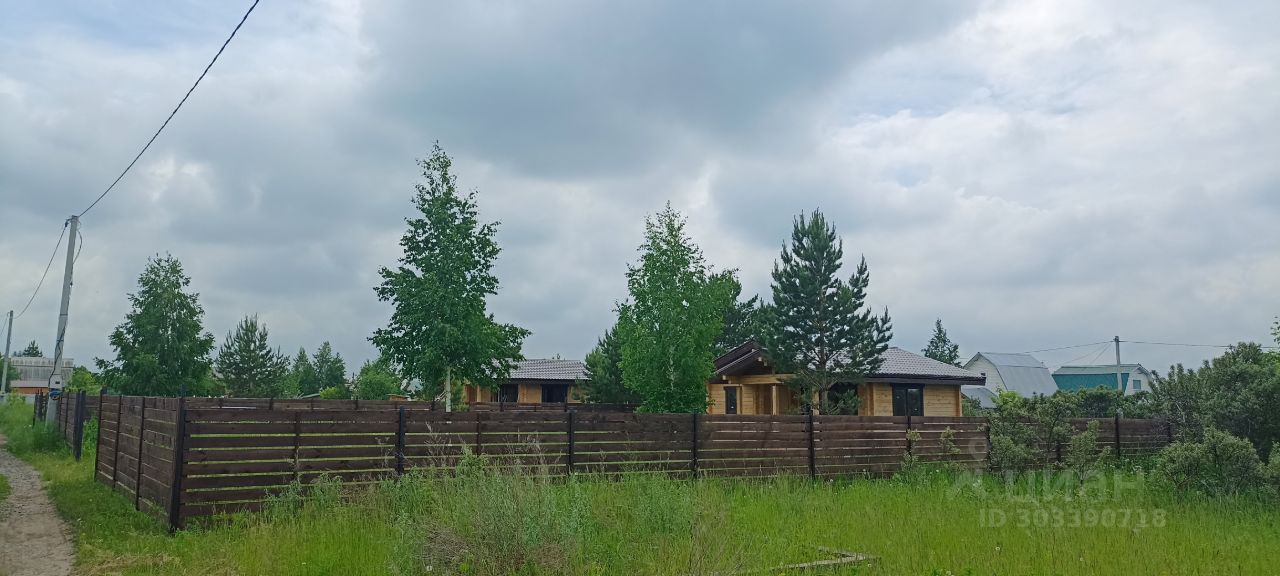 На фотографии изображены деревянные дома в загородном комплексе, выполненные в традиционном стиле. Структурные элементы включают крыши с наклоном и большие окна, что создает светлую и уютную атмосферу. Дома окружены высоким забором из деревянных панелей, который обеспечивает уединение. Вокруг виднеются зеленые деревья и кустарники, что подчеркивает природный ландшафт. Основные цвета — натуральные деревянные оттенки и зеленый, что создает гармоничное сочетание с окружающей природой. Используемые материалы придают постройкам экологичный и привлекательный вид.