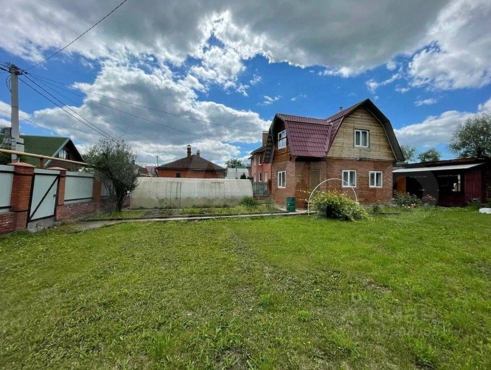 Двухэтажный дом в стиле загородной архитектуры с деревянными и кирпичными элементами. Крыша покрыта черепицей, окна имеют простую форму. Видно, что дом окружён ухоженным участком с зелёной травой и небольшими кустарниками. Внешние стены выполнены из кирпича и дерева, что придаёт дому уютный вид. На участке присутствует небольшой забор, дополненный воротами. Пространство выглядит открытым и просторным, что создаёт атмосферу уединения и спокойствия.