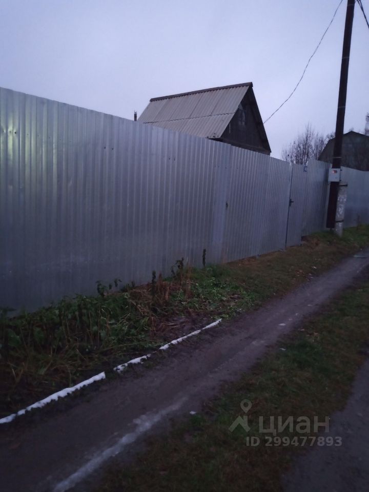 На фотографии виден металлический забор, который окружает участок. Забор выполнен из гофрированного металла, что придаёт ему современный вид и обеспечивает приватность. На заднем плане можно заметить одноэтажное строение с крышей из металлочерепицы, имеющее характерный для дачных участков дизайн. Около забора растут травы и кустарники, что свидетельствует о том, что территория не полностью ухожена. Дорога, проходящая рядом, имеет грунтовое покрытие и обрамлена белой линией, что добавляет контраст к общей картине. Освещение на фотографии тусклое, создающее атмосферу спокойствия.
