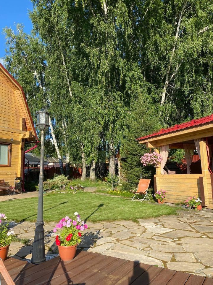 На фото представлен загородный участок в русском стиле с деревянными строениями. Архитектурные элементы включают крыши с красной черепицей и деревянные панели. Видно ухоженное зеленое газонное пространство, окруженное цветочными клумбами в горшках с яркими цветами. Структурные особенности включают беседку с занавесками и удобным креслом, а также фонарь, создающий атмосферу уюта. На каменной дорожке видны большие плиты, гармонично сочетающиеся с природными материалами. Цветовая палитра включает теплые древесные оттенки и яркие цветы, создавая приятное визуальное восприятие.