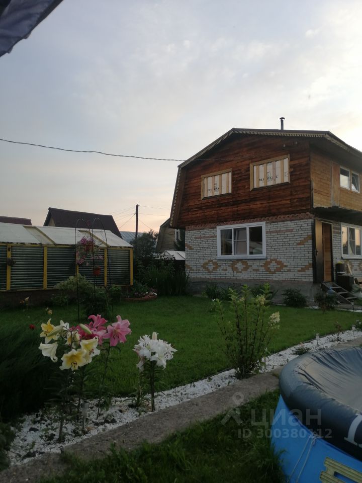 Двухэтажный дом в загородном стиле с деревянным фасадом и кирпичными элементами. На первом этаже расположены большие окна, обеспечивающие естественное освещение. Внешний вид включает аккуратный газон с цветами, среди которых выделяются лилии, и декоративные камни, создающие ухоженный вид. Структурные элементы включают террасу у входа и балкон на втором этаже. Цветовая гамма здания сочетает натуральные древесные оттенки с белыми и кирпичными элементами, что придаёт дому привлекательный и гармоничный вид. Пространство вокруг оформлено с заботой о ландшафтном дизайне.
