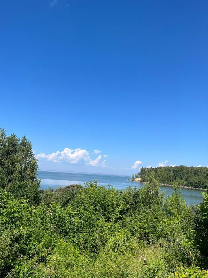 На изображении виден просторный водоем с гладкой поверхностью, отражающей небесные облака. Береговая линия окружена густой зеленью, включая кустарники и деревья, создающими естественный ландшафт. Вода имеет спокойный, прозрачный оттенок, а небо ярко-синее с легкими облаками. Пейзаж выглядит умиротворяющим и живописным, подчеркивая красоту природы. Элементы природы гармонично сочетаются, создавая ощущение уединения и спокойствия.