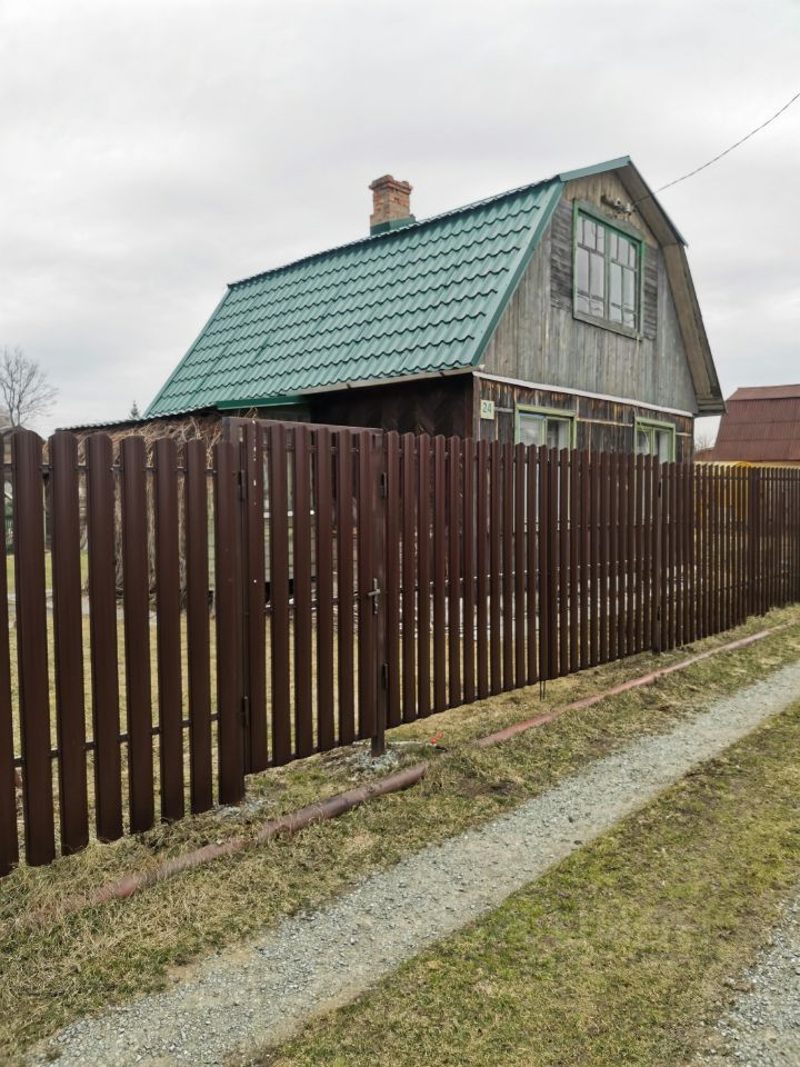 Деревянный дом в загородном стиле с крышей из зеленой металлочерепицы. Фасад отделан темным деревом, что придает ему уютный вид. Окна с двойными рамами и декоративными элементами создают атмосферу традиционного русского зодчества. Забор из деревянных планок коричневого цвета обрамляет участок, обеспечивая приватность. Входная дверь расположена в центре фасада, что делает акцент на симметрии. Общая композиция дома гармонично вписывается в загородный ландшафт, создавая ощущение спокойствия и уюта.